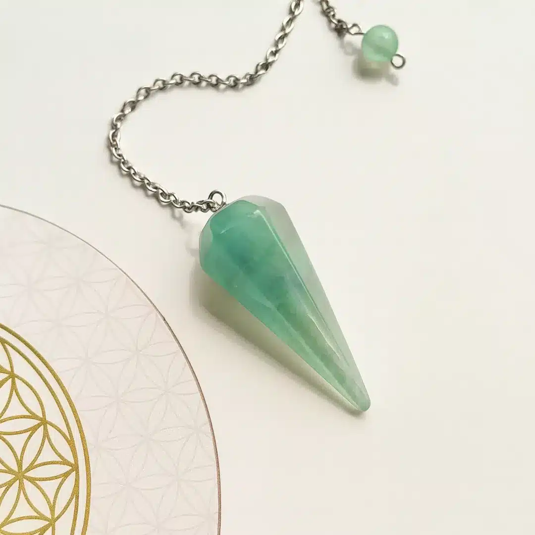 Pendule divinatoire conique en fluorite naturelle avec chaîne argentée, posé sur une fleur de vie dorée, idéal pour la radiesthésie et l’intuition.
